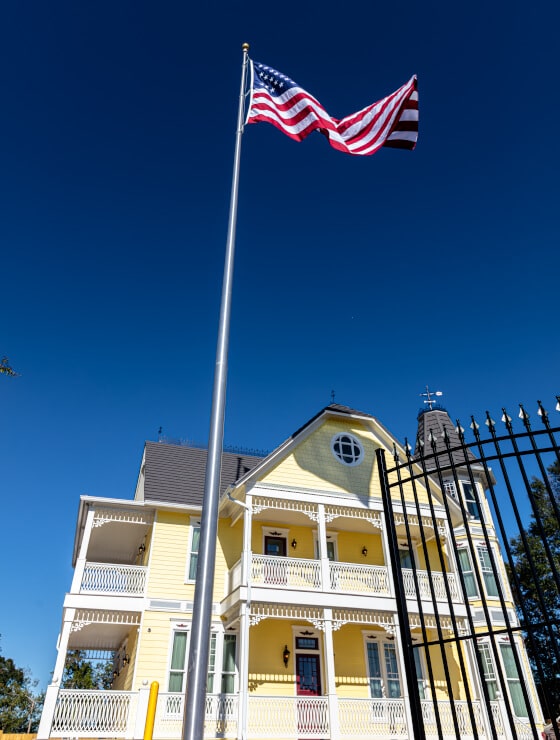 An angled picture of a multi story yellow Victorian house with white trim, a wrought iron fence, and an American flag out front.
